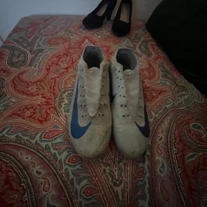 Nike vita fotbollsskor med blå detaljer - Säljer ett par vita Nike fotbollsskor med blå swoosh och detaljer. Skorna har låg profil, platt sula med dobbar och är tillverkade i syntetmaterial. Perfekta för dig som vill ha stilrena och sportiga skor på planen.