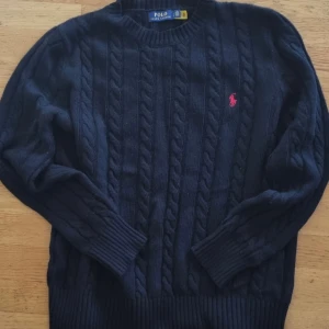 Svart kabelstickad tröja Polo Ralph Lauren - Tröjan är i helt nyskick, knappt använd eftersom den var lite för stor för mig. Kabelstickad svart tröja från Polo Ralph Lauren med klassisk rund halsringning och röd broderad logga på bröstet. Tröjan är långärmad och har ribbade muddar samt nederkant. Perfekt för dig som gillar stilren och tidlös design samt vill hålla dig varm.