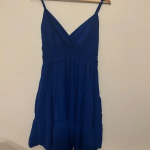 Blå kort klänning med volanger - Supersöt blå klänning med smala axelband och v-ringad omlott framtill. Klänningen har smock i midjan och flera volanglager som ger en flowy känsla. Perfekt för varma dagar och somriga vibes.