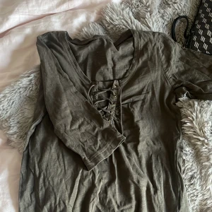 Olivgrön topp med snörning fram - Snygg olivgrön topp med trekvartsärm och cool snörningsdetalj framtill. Toppen har en rundad halsringning och är tillverkad i ett mjukt, lätt material som känns skönt mot huden. Perfekt för dig som gillar en avslappnad men trendig stil.