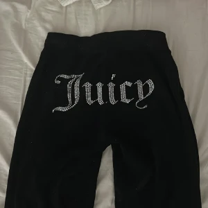 Svarta Juicy Couture mjukisbyxor XXS - Svarta mjukisbyxor från Juicy Couture i storlek XXS. Byxorna har snörning i midjan och är dekorerade med strass som bildar texten 'Juicy' på baksidan. Materialet är mjukt och har en lyxig känsla, perfekt för en chill och trendig look.