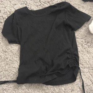 Svart ribbad croppad topp med snörning - Säljer en svart, ribbad croppad topp med korta ärmar och snörningsdetalj i sidan. Toppen har rund halsringning och är i ett mjukt, stretchigt material som sitter snyggt på kroppen. Perfekt för dig som gillar en enkel men trendig stil.