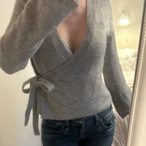 Grå omlottkofta med knyt i sidan - Supermysig grå omlottkofta med djup v-ringning och knyt i sidan. Koftan är stickad och har långa ärmar med lite lösare passform. Perfekt att slänga på sig för en chill och snygg look.