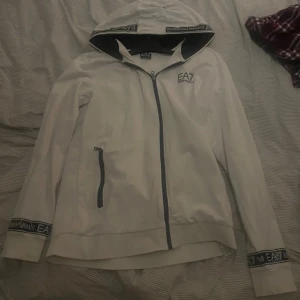 Vit hoodiejacka från Emporio Armani EA7 - Vit hoodiejacka med svarta detaljer och dragkedja från Emporio Armani EA7. Jackan har huva, muddar med logotyp och en ficka med dragkedja på ena sidan. Perfekt för en sportig och avslappnad stil.