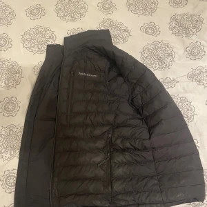 Svart dunväst från Peak Performance - Svart dunväst från Peak Performance med quiltad design och hög krage. Västen har dragkedja framtill, två sidofickor med dragkedja och en bröstficka. Perfekt för lager-på-lager och outdoor-vibe. Storlek m-l