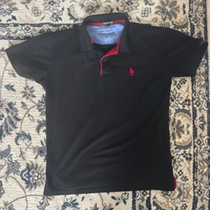 Svart pikétröja från Polo Ralph Lauren - Svart pikétröja från Polo med klassisk krage och knäppning framtill. Röd broderad logga på bröstet och röda detaljer vid knappar och insida krage. Tillverkad i bomull för en skön känsla. Perfekt för en clean och stilren look.