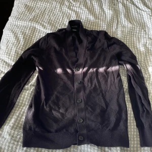 Svart merinoullskofta från Riley - Mörk, klassisk svart kofta från Riley i mjuk merinoull. Koftan har lång ärm, djup v-ringning och knäpps med sex knappar framtill. Perfekt för lager-på-lager och enkel att styla till olika looks.