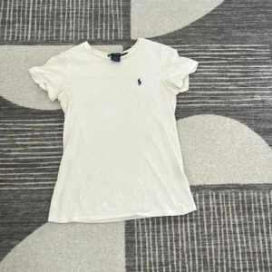 Vit t-shirt från Ralph Lauren - Klassisk vit t-shirt från Ralph Lauren med liten mörkblå broderad logga på bröstet. T-shirten har rund halsringning och korta ärmar. Tillverkad i mjuk bomull för en skön och avslappnad känsla. Perfekt basplagg till garderoben.