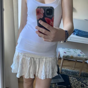 Vit blommig kortkjol med volang - Supersöt vit kortkjol med små blommor i pastellfärger och bred smock i midjan. Kjolen har volang nertill som ger en flowy känsla och är gjord i ett lätt och luftigt material, perfekt för varma dagar. Högmidjad men går att vika mer så att den blir kortare.💕 lappen är kvar men klippt för att det var en present. 💕💕