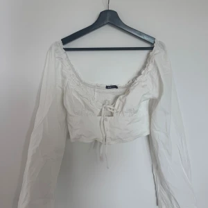 Vit croppad blus från Gina Tricot - Säljer en söt vit croppad blus från Gina Tricot med långa ärmar, smockad överdel och knytning framtill. Blusen har en fyrkantig ringning med volangdetaljer och knappar framtill. 
