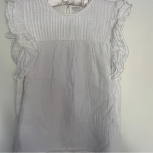 Vit volangblus från Lindex - Supersöt vit blus från Lindex med volangdetaljer på ärmarna och rynkad struktur upptill. Blusen är ärmlös med rund halsringning och har en luftig känsla, perfekt för varma dagar. Tillverkad i mjukt bomullstyg.