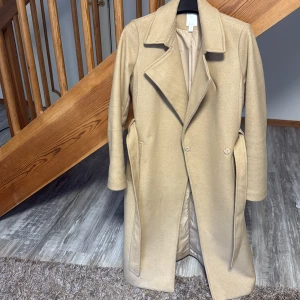 Beige kappa från H&M med bälte - Snygg beige kappa från H&M i storlek 38. Klassisk modell med bred krage, knappar framtill och bälte i midjan. Långärmad och med fickor på sidorna. Perfekt för dig som gillar stilrena och tidlösa plagg.