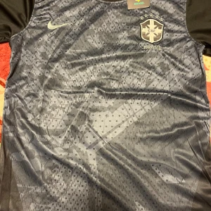 Svart Brasil Nike fotbollströja - Snygg svart Brasilien fotbollströja från Nike med silverdetaljer och CBF-emblem på bröstet. Tröjan har korta ärmar, rund hals med ränder och är tillverkad i lätt, ventilerande Dri-FIT-material. Unikt mönster över hela framsidan för extra stil.