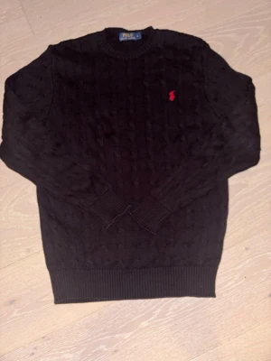 Svart kabelstickad tröja Polo Ralph Lauren - Kabelstickad svart tröja från Polo Ralph Lauren med klassisk röd logga broderad på bröstet. Tröjan har rund halsringning, ribbade muddar och nederkant. Perfekt för dig som gillar stilrena och tidlösa plagg.
