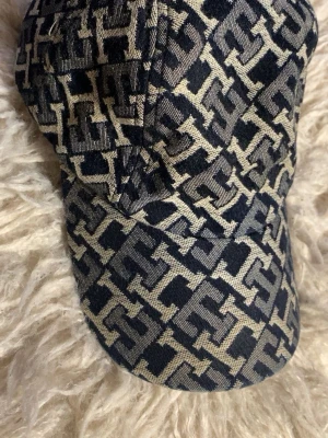 Mönstrad keps från Tommy Hilfiger - Snygg keps i mörkblått och beige med all-over TH-monogram från Tommy Hilfiger. Klassisk böjd skärm och justerbar rem baktill med metallspänne. Tillverkad i ett vävt tyg som ger en exklusiv känsla och cool look.