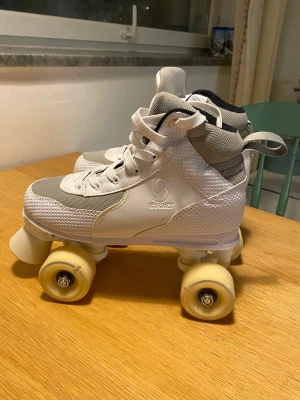 Vita rullskridskor från Story - Snygga vita rullskridskor från Story med högt skaft, vita snören och meshdetaljer i grått. Skorna har robusta hjul i cremefärg och tåskydd framtill. Perfekta för dig som vill rulla runt med stil och komfort. Aldrig använda! 