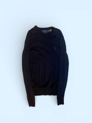 Mörkblå tröja från Polo Ralph Lauren - Klassisk mörkblå tröja från Polo Ralph Lauren i mjuk merinoull. Tröjan har rund halsringning, diskret broderad logga på bröstet och ribbade muddar vid ärmslut och nederkant. Perfekt för en stilren look.OBS den här några små hål men de går så klart att laga.