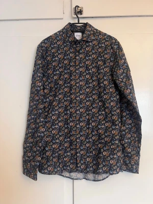 Blommig skjorta från Oscar of Sweden - Snygg slim fit skjorta från Oscar of Sweden med mörkblå bas och blommigt mönster i blått, brunt och vitt. Skjortan har klassisk krage och långa ärmar. Perfekt för dig som gillar att sticka ut med stilrena detaljer.