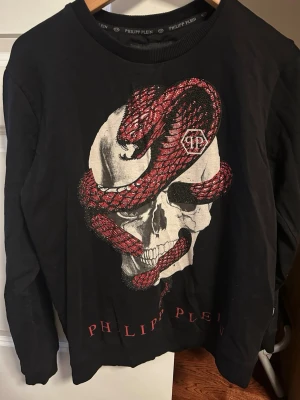 Svart långärmad tröja från Philipp Plein - Svart långärmad topp från Philipp Plein med stor dödskalle och röd orm framtill. Rund halsringning och logotypdetalj i nacken. Snygg och edgy design med tryck i vitt, rött och svart. Perfekt för dig som gillar statement-plagg.