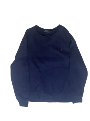 Mörkblå sweatshirt från Polo Ralph Lauren - Klassisk mörkblå sweatshirt från Polo Ralph Lauren med rund halsringning och röd broderad logga på bröstet. Tröjan har långa ärmar och ribbade muddar vid ärmslut och nederkant. Perfekt för en avslappnad och stilren look.
