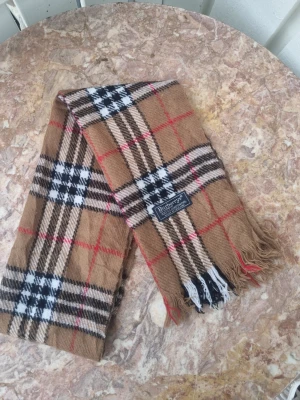 Burberrys beige rutig halsduk ull - Klassisk beige halsduk från Burberry i 100% lammull med ikoniskt rutmönster i svart, vitt och rött. Halsduken har fransar i ändarna och en vävd etikett med Burberrys-loggan. Perfekt accessoar för att lyfta din outfit.