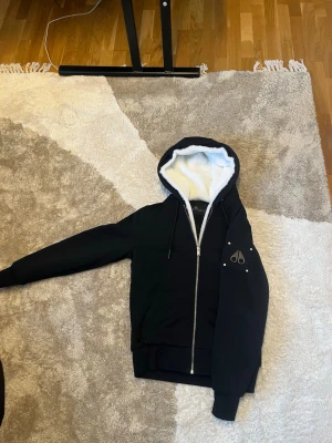 Svart jacka med dragkedja och huva - Svart hoodie från Moose Knuckles med vit fluffig insida i huvan och dragkedja framtill. Går att ta av pälsen på huvan och det är en äkta vinterjacka köpt på johnells. Den är xs men passar S 