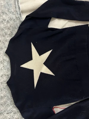 Mörkblå tröja med vit stjärna från Gina tricot - Tröja från Gina tricot. Mörkblå tröja med en stor vit stjärna på framsidan. Tröjan har rund halsringning och långa ärmar. Materialet är mjukt och stretchigt.