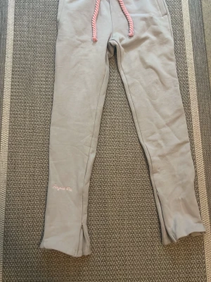 Grå mjukisbyxor med rosa detaljer - Snygga grå mjukisbyxor från Frejuse Vibe med rosa snörning i midjan och coolt rosa tryck bak. Byxorna har slits vid benslut och är perfekta för en avslappnad stil. Mjukt material som känns skönt mot huden.