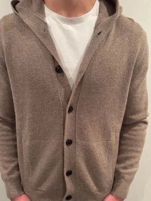 Cardigan - Mysig beige kofta med huva och svarta knappar framtill. Tröjan har två fickor fram och ribbade muddar vid ärmslut och nederkant. Perfekt för lager på lager och chill stil. Materialet ser ut att vara mjukt och stickat, troligen bomull eller ullmix.