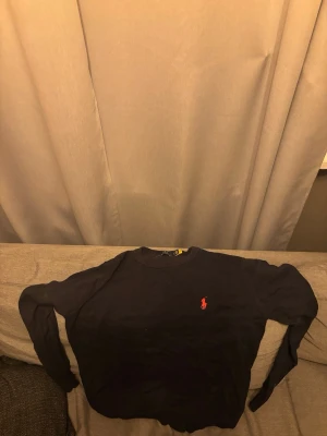 Mörkblå tröja från Ralph Lauren - Klassisk mörkblå tröja från Ralph Lauren med den ikoniska röda loggan broderad på bröstet. Tröjan har rund halsringning och långa ärmar, perfekt för en stilren och enkel look.