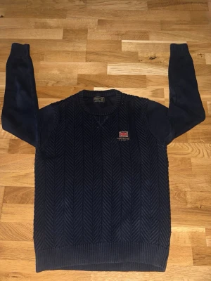Mörkblå stickad tröja med mönster från Morris - Mörkblå stickad tröja från Gant med fiskbensmönster och rund hals. Tröjan har en liten broderad patch med brittiska flaggan och Gant-loggan på bröstet. Ribbstickade muddar vid ärmslut och nederkant. Perfekt för kyliga dagar.