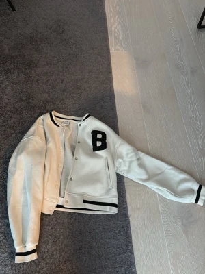 Zara tjocktröja/jacka - Snygg vit bomberjacka från JUNKYARD med svarta detaljer, broderad B på bröstet och texten Brooklyn 82 på ryggen. Jackan har klassisk basebollstil med ribbade muddar och krage samt tryckknappar framtill. Perfekt för en sportig och trendig look.
