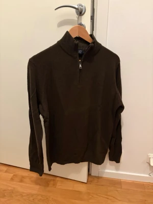 Brun stickad tröja från Hackett - Mörkbrun stickad tröja från Hackett London i 100% lammull. Tröjan har hög krage med halv dragkedja och ribbstickade detaljer vid halsen. Perfekt för dig som gillar klassisk och stilren design.