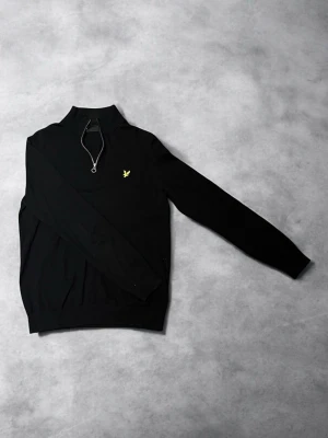 Svart halvzip-tröja från Lyle & Scott - Svart långärmad  tröja från Lyle & Scott i mjukt material med halv dragkedja och gul broderad logga på bröstet. Tröjan har ribbade muddar och hög krage, perfekt för en clean och stilren look. Materialet känns mjukt och bekvämt.
