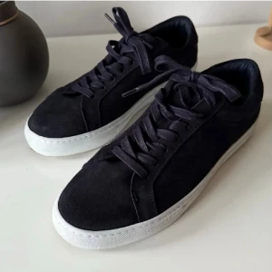 Marinblåa sneakers från Les Deux - Stilrena marinblå sneakers från Les Deux i mocka med vita sulor och svarta skosnören. Diskret detalj med små ränder i rött, vitt och blått på hälen. Perfekta för en clean och modern look.