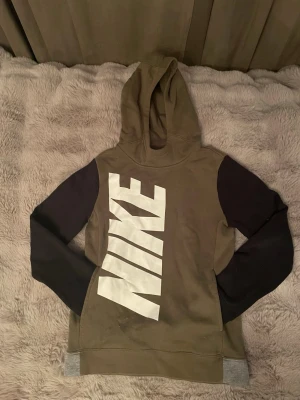 Nike hoodie med stora loggan - Snygg hoodie från Nike i olivgrönt med svarta ärmar och gråa muddar. Stor vit Nike-logga tryckt diagonalt över framsidan. Tillverkad i mjukt bomullsmaterial och har en klassisk huva. Perfekt för en sportig och avslappnad stil. (Det är en barn hoddie men måste ta m för fanns inte mycket annat att välja på)