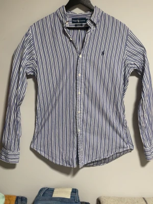 Randig skjorta från Ralph Lauren - Snygg skjorta från Ralph Lauren i vitt och blått med klassiska vertikala ränder. Modellen är Custom Fit och har lång ärm, knäppning framtill. Perfekt för en stilren look.