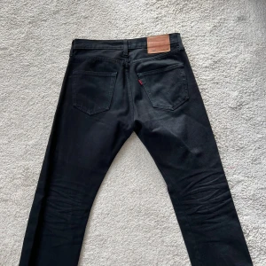 Svarta Levi's 501 jeans storlek W30 L30 - Säljer ett par klassiska svarta Levi's 501 jeans med raka ben och knappgylf. Jeansen har fem fickor, kopparfärgade nitar och den ikoniska läderpatchen bak i midjan. Perfekta för en clean och tidlös look.