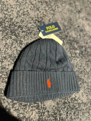 Mörkgrå kabelstickad mössa Polo - Snygg mörkgrå kabelstickad mössa från Polo Ralph Lauren med uppvikt kant och klassisk orange logga framtill. Tillverkad i mjukt material som håller dig varm och stilsäker under kalla dagar.