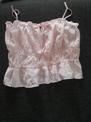 Ljusrosa volangig crop top - Söt ljusrosa crop top med tunna axelband och volangkant nedtill. Toppen har en liten rosett framtill och smockdetaljer som ger en avslappnad och feminin look. Perfekt för sommardagar och enkel att matcha med jeans eller kjol.