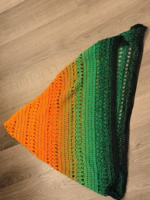 Virkad trekantssjal i orange och grönt - Handvirkad triangelsjal i snygg färgskiftning från orange till grönt och svart. Virkat med Halloween Dahlia i 100% bomullsgarn. Sjalen har ett luftigt mönster med hål och detaljerade virkade partier. Pris är för material + tidsfördriv.