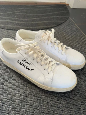 Vita Saint Laurent sneakers - Snygga vita sneakers från Saint Laurent med handskriven logga på sidan. Skorna är i läder med vita snören och klassisk låg profil. Perfekta för dig som gillar stilrena och exklusiva sneakers med en unik touch.