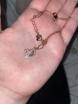 Swarvoski Roséguldfärgat halsband med svanberlock - Säljer ett elegant halsband i roséguld med en svanformad berlock dekorerad med små gnistrande stenar och en större pärlliknande detalj i mitten. Kedjan är tunn och smidig, perfekt för att ge en stilren touch till din outfit.