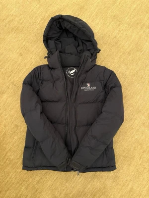 Svart dunjacka från Kingsland - Svart dunjacka från Kingsland med huva och dragkedja framtill. Jackan har två sidofickor med dragkedja och broderad logga på bröstet. Perfekt för kalla vinterdagar och riktigt skön att bära.