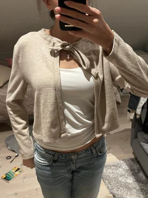 Beige cropped kofta med knytning - Snygg beige cropped kofta med knytning vid halsen och långa ärmar. Perfekt att slänga över ett linne eller t-shirt för en chill och trendig look. Mjuk och lätt material som känns skönt mot huden.