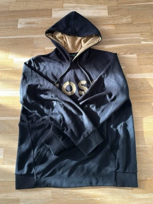 Svart hoodie från BOSS med guldtext - Snygg svart hoodie från BOSS med stor guldfärgad logga på bröstet. Hoodien har en beige insida i huvan och svarta dragsnören. Tillverkad i mjukt bomullsmaterial och har ribbade muddar vid ärmslut och nederkant. Perfekt för en avslappnad och trendig look.