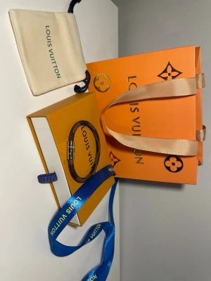 Louis Vuitton brunt armband - Snyggt armband från Louis Vuitton i brunt canvasmaterial med klassiskt monogrammönster och ett silverfärgat magnetlås med ingraverad logga. Kommer med originalförpackning, dustbag och påse. Perfekt accessoar för att lyfta din stil.
