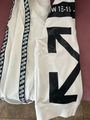 Vita mjukisdress  - Snygga vita mjukisdressfrån med svarta grafiska tryck längs ena benet och logoband på sidorna. Byxorna har resår i midjan och vid bensluten för en skön passform. Perfekta för en streetwear-look.