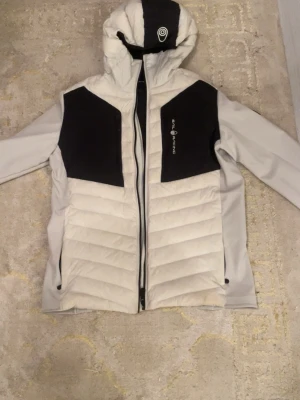 Vit och svart dunjacka från Sail racing - Snygg dunjacka från Sail racing i vitt och svart med huva och dragkedja framtill. Jackan har svarta partier på bröstet och huvan, samt diskret logga på bröstet. Insidan är svart och jackan har quiltad design för extra värme. Perfekt för kalla vinterdagar.