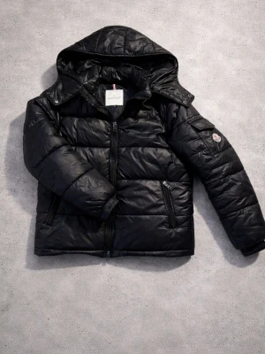 Svart dunjacka från Moncler med huva - Snygg svart dunjacka från Moncler med huva och dragkedja framtill. Jackan har två sidofickor med dragkedja och en Moncler-logga på ärmen. Perfekt för kalla vinterdagar och riktigt stilren look.